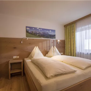 A cozy bedroom with a double bed and wooden paneling. The room features large windows with curtains and a harmonious decoration.