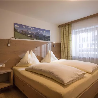 A cozy bedroom with a double bed and two large pillows. A picture of the mountains hangs on the wall, while the window lets in plenty of daylight.