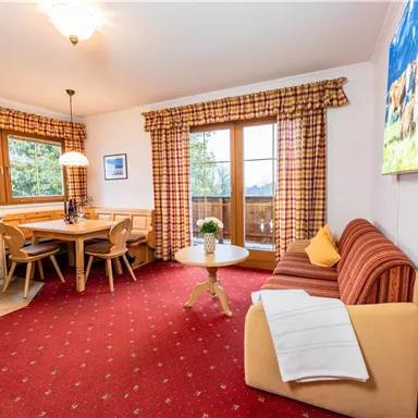 Ein gemütliches Wohn- und Esszimmer mit einem Esstisch, einem Sofa und großen Fenstern. Die Einrichtung ist hell und freundlich, mit einem roten Teppich und bunten Vorhängen.