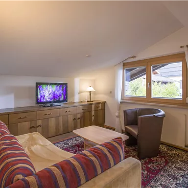 A cozy living room with a sofa, a table, and a television. Large windows let in plenty of daylight.