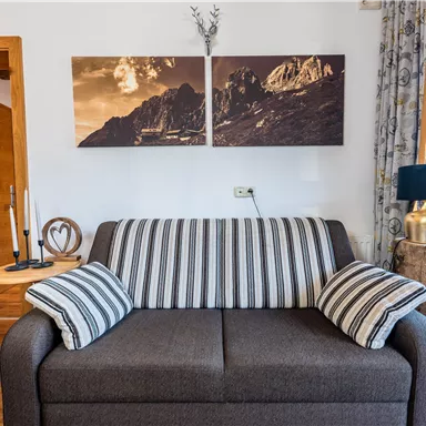 Ein gemütliches Wohnzimmer mit einem grauen Sofa und gestreiften Kissen. An der Wand hängen zwei Bilder von Bergen und ein kleiner Tisch steht neben dem Sofa.