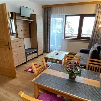 Ein gemütlicher Wohnraum mit Holzfußboden und modernen Möbeln. Es gibt einen Esstisch mit Blumen, einen Fernseher und große Fenster, die viel Licht hereinlassen.