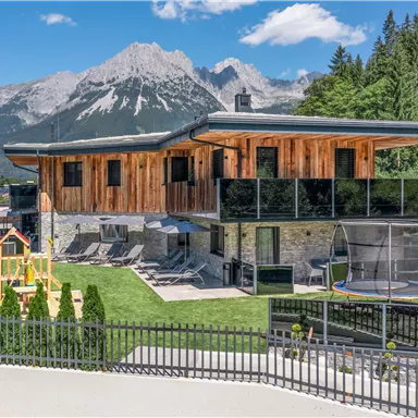 A modern wooden house with large windows situated in a mountainous landscape. In the garden, there is a swing and a trampoline.