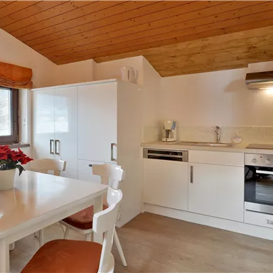 Eine moderne Küche mit einem Esstisch und weißen Möbeln. Der Boden ist hell, und das Holzdecke sorgt für eine warme Atmosphäre.