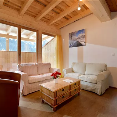 Ein gemütliches Wohnzimmer mit Holzdecke und großen Fenstern. Es gibt bequeme Sofas und einen Holztisch mit einem Blumenarrangement.
