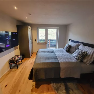 A modern bedroom with a large bed and decorative pillows. A television is mounted on the wall, and there is a window that lets in natural light.