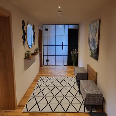 A bright hallway with wooden flooring and modern design. A mirror and a picture are hanging on the wall, and there are two seating areas on a geometric rug.