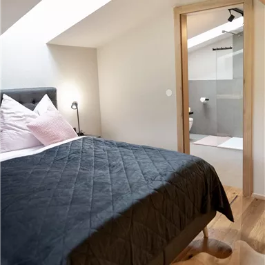 A modern bedroom with a comfortable bed and soft color tones. Behind it is a bathroom with elegant tiles.