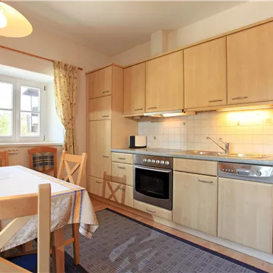A bright kitchen with wooden furniture and modern equipment. On the table is a place setting, and the window lets in daylight.
