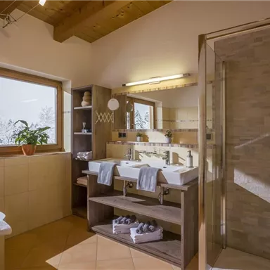 A modern bathroom with two sinks and a large shower. Bright tiles and a window bring in plenty of light.