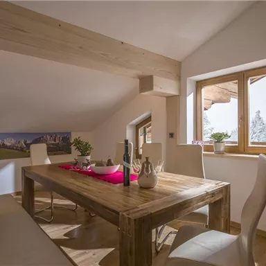 A cozy dining area with a large wooden table and white chairs. Through the window, you can see a snowy landscape.