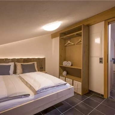 A cozy bedroom with a double bed, wood details, and modern furnishings. The room is well-lit and has access to another area.