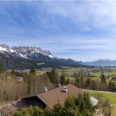 A picturesque landscape with snow-covered mountains and green meadows. In the foreground, there is a beautiful house and trees visible.