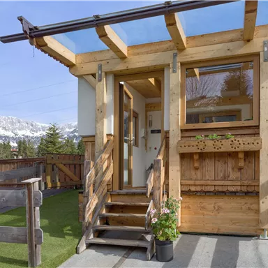 A charming wooden house with an inviting front door and a porch. In the background, mountains and a clear sky can be seen.