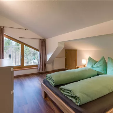 A cozy bedroom with two beds and light green bedding. Large windows let in plenty of daylight and offer a view of the greenery.