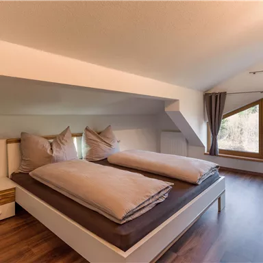 A cozy bedroom with a double bed and large windows. The room has a modern design and a warm wooden floor.
