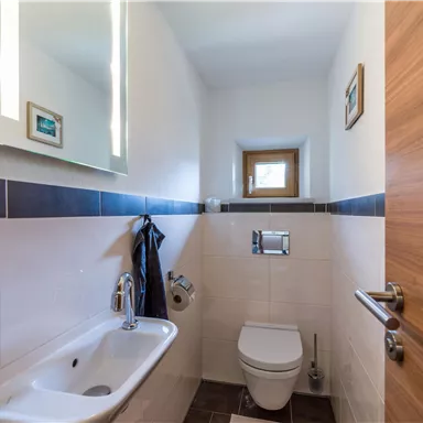 A modern bathroom with a toilet, a washbasin, and a mirror. The walls are light-colored, and there is a small window for natural light.