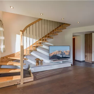 A modern living room with a wooden staircase and a large television. The walls are painted light and there is natural light coming through the windows.