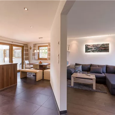 A modern living room with a cozy sofa and a table. The open space offers a view of the kitchen and has large windows that let in daylight.