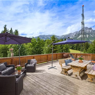 A cozy terrace with seating and sun umbrellas. In the background, forests and mountains can be seen under a blue sky.