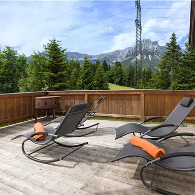 A cozy balcony with lounge chairs and orange towels. In the background, tall trees and the mountains can be seen.
