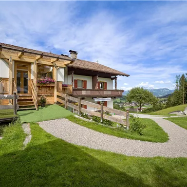A rustic house in the midst of nature with green grass. In the background, mountains and a clear sky can be seen.