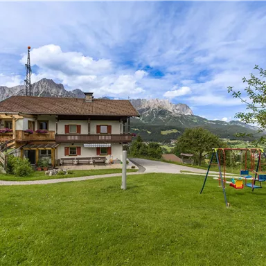 A cozy house in a green landscape with a playground. In the background, majestic mountains and a blue sky are visible.