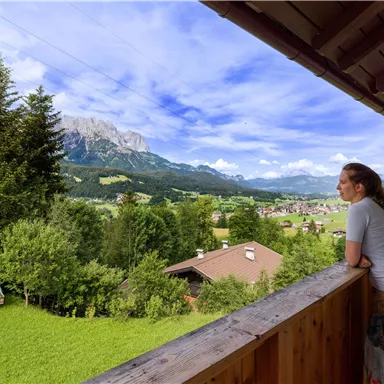 A woman stands on a balcony and looks at the beautiful mountain landscape. In the background, green meadows and forested hills can be seen.