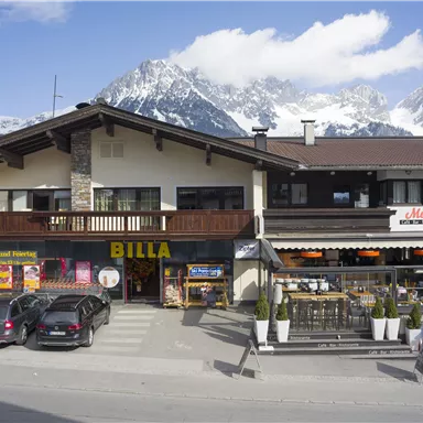 A commercial building in the mountains with the "BILLA" logo on the ground floor. In the foreground, parked cars and a terrace with tables and chairs are visible.
