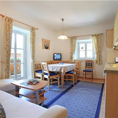 A bright living room with a dining table and chairs. The kitchen is well-equipped, and there are large windows that let in a lot of light.