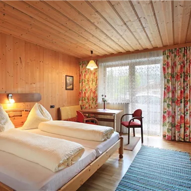 A cozy room with wood paneling, two beds, and a small table. Daylight shines through the window, and the curtains are decorated with colorful floral patterns.