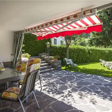 Eine einladende Terrasse mit einem Essbereich und einem Sonnenschirm. Der Garten ist grün mit vielen Sitzgelegenheiten im Freien.