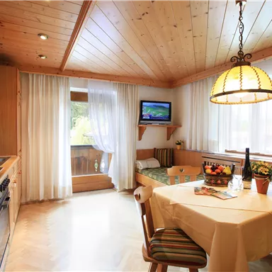 A cozy kitchen with wooden paneling and a table set with fruit and a bottle of wine. A small television hangs on the wall and bright curtains let in plenty of light.