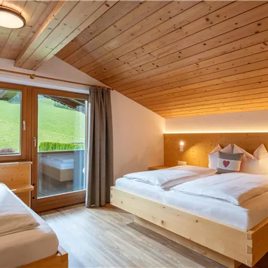 Ein gemütliches Zimmer mit Holzdecke, zwei Betten und großen Fenstern. Draußen ist eine grüne Wiese zu sehen.