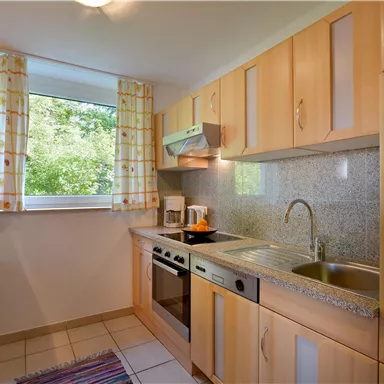 A modern kitchen with wooden cabinets and a window overlooking the greenery. It is equipped with a stove, a sink, and a coffee machine.