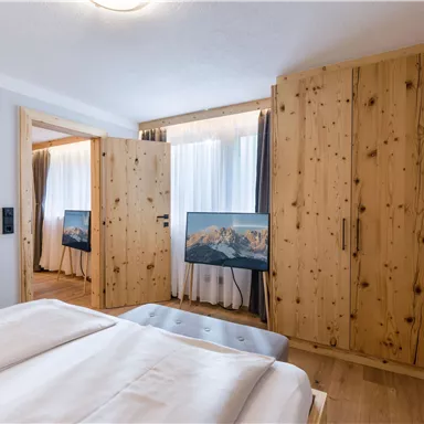 A modern bedroom with a wooden wardrobe and two televisions. The windows are decorated with curtains and let in plenty of daylight.