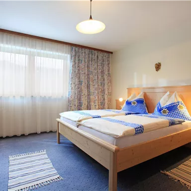 A cozy bedroom with a double bed and blue pillows. Large windows let in plenty of daylight and the furniture is made of wood.