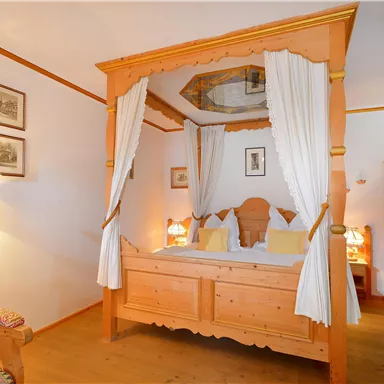 Ein gemütliches Schlafzimmer mit einem vierteiligen Bett aus Holz und weißen Vorhängen. Das Zimmer hat warmes Licht und traditionelle Dekorationselemente.