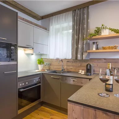 Eine moderne Küche mit hellen Schränken und Holzdetails. Auf der Arbeitsplatte stehen zwei Weingläser und eine Flasche Wein.