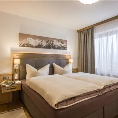 Ein gemütliches Zimmer mit einem großen Doppelbett und stilvoller Holzdekor. Große Fenster sorgen für viel Tageslicht und einen Blick auf die Berge.