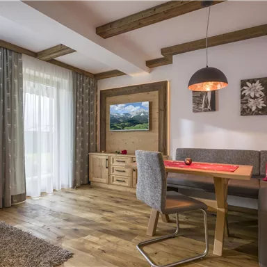 Ein modernes Wohnzimmer mit Holzdecke und einem Essbereich. Die Fenster bieten viel natürliches Licht und die Dekoration ist gemütlich und einladend.