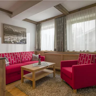 Ein gemütliches Wohnzimmer mit roten Sofas und einem Holztisch. Große Fenster sorgen für viel Tageslicht und eine einladende Atmosphäre.
