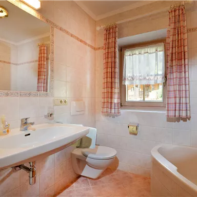A bright bathroom with a sink, a toilet, and a bathtub. The walls are tiled and there are curtains at the windows.