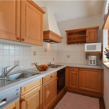 A modern kitchen with wooden cabinets and a bright work surface. There is a fruit bowl on the countertop, and the room has a window with curtains.