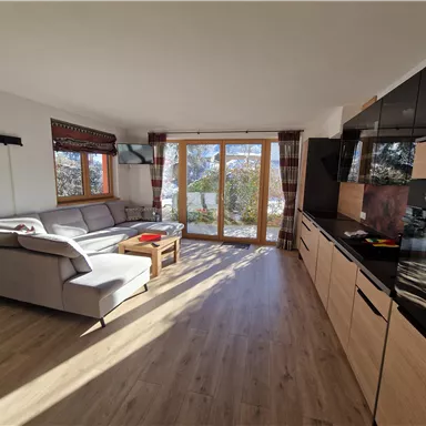 Ein modernes Wohnzimmer mit einem großen Sofa und einem Holztisch. Große Fenster lassen viel Tageslicht herein und bieten einen schönen Blick nach draußen.