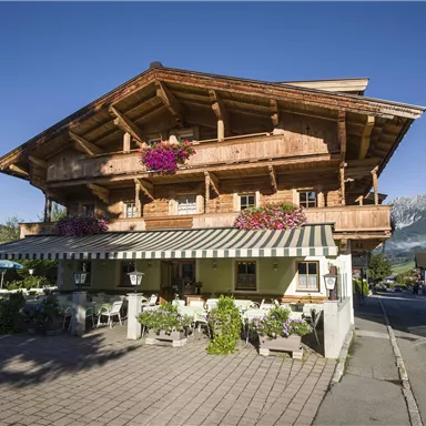 Ein traditionelles Alpenhaus mit Holzverzierungen und blühenden Pflanzen. Die Aussicht auf die umliegenden Berge ist klar und einladend.