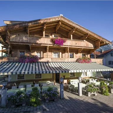 Ein traditionelles Holzhaus in den Alpen mit blühenden Balkonkästen. Vor dem Gebäude befinden sich gemütliche Sitzgelegenheiten im Freien.