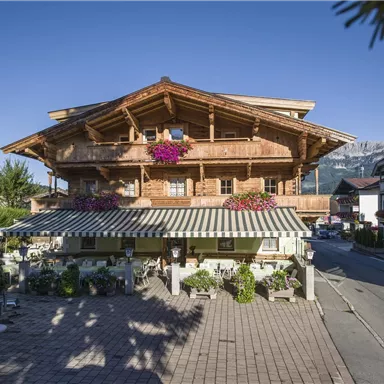 Ein traditionelles Holzhaus mit Blumen und einem schönen Balkon. Im Vordergrund sieht man ein gemütliches Café und eine malerische Straße mit Bäumen.