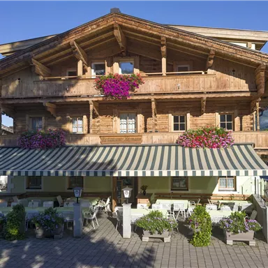 Ein traditionelles Holzhaus mit einem großen Balkon und bunten Blumen. Vor dem Haus befindet sich ein einladendes Gasthaus mit Terrassenmöbeln.