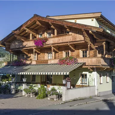 Ein traditionelles, zweigeschossiges Holzhaus mit Blumenschmuck. Umgeben von Bäumen und einer klaren Berglandschaft.